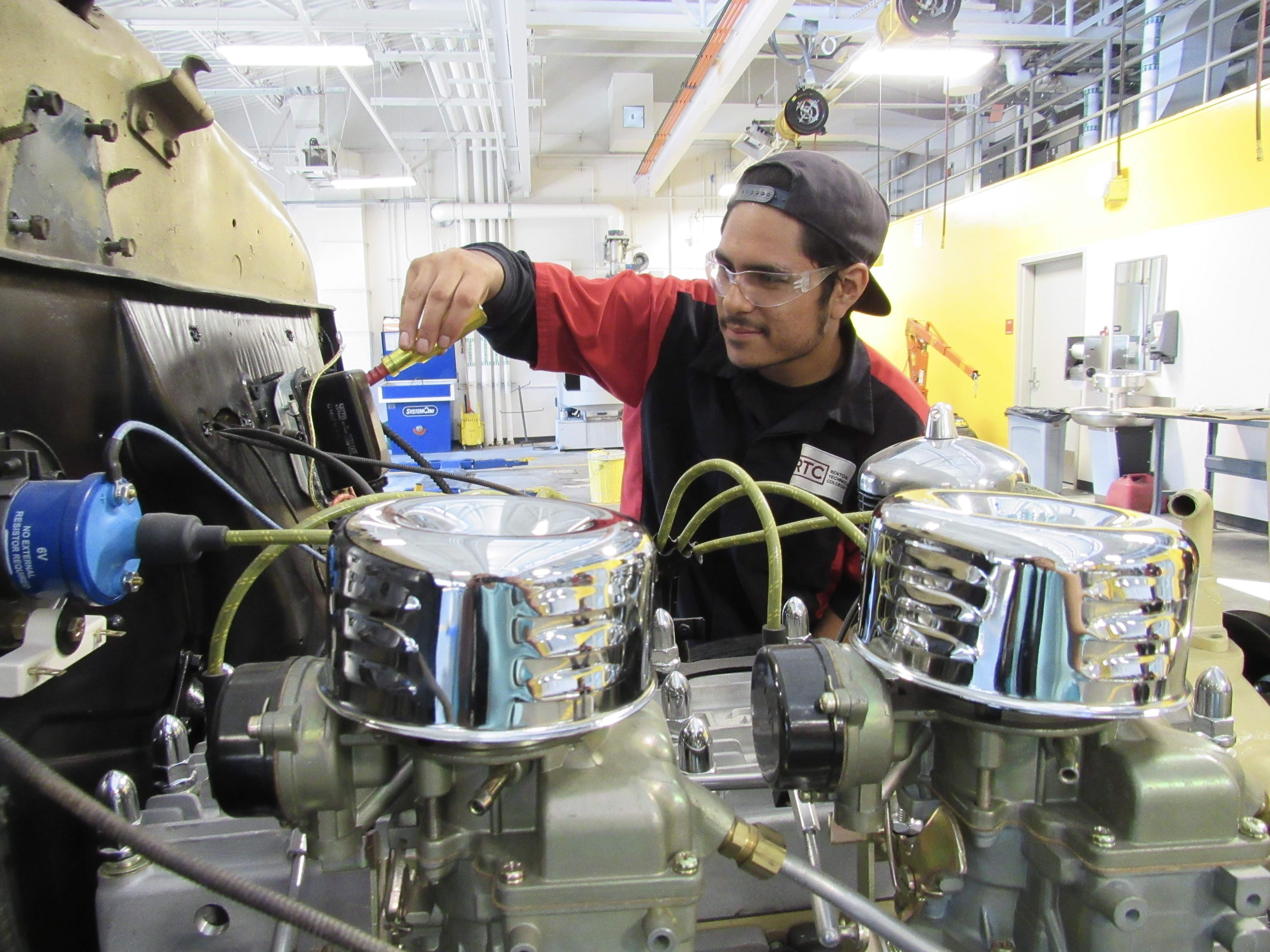 Two students interacting with a large industrial machine using a red button, and both appear engaged and smiling. They are wearing casual work attire and safety glasses.